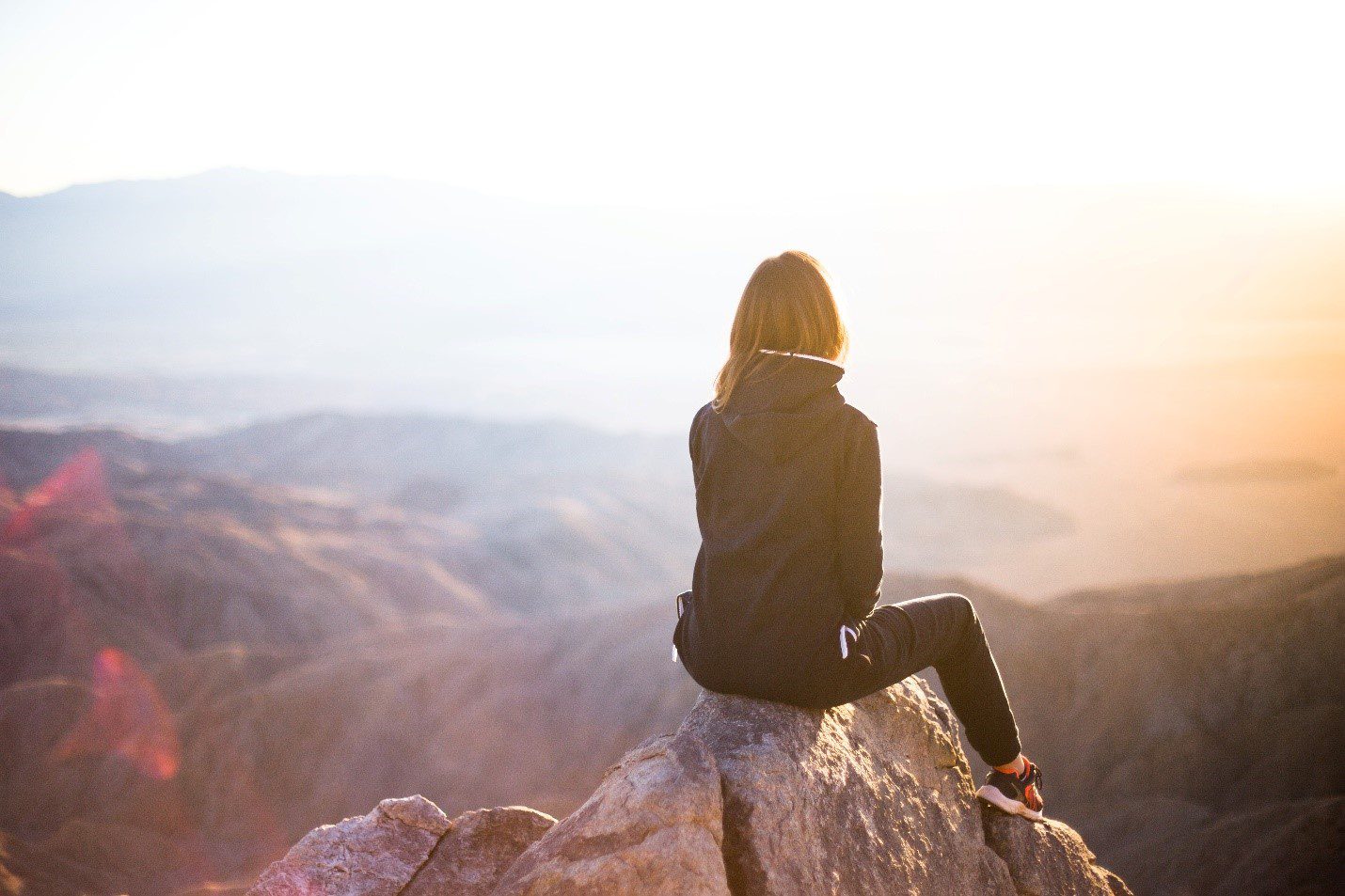 A woman with her back to the camera sits atop a rock and looks out over a mountain range.