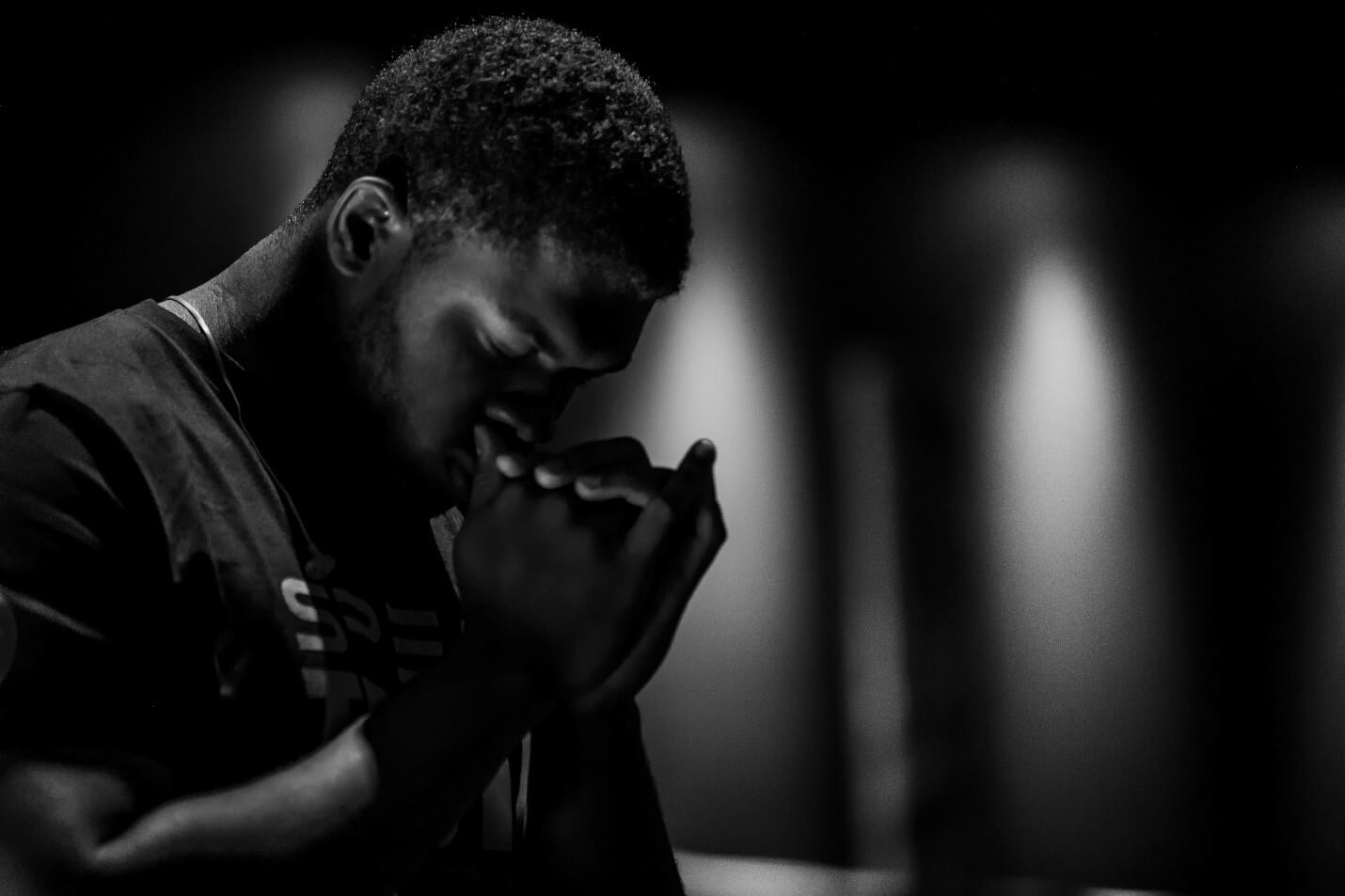 A Black man with his hands clasped in front of his face with his head bowed in a prayer pose.