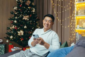 Portrait of man holding phone for Christmas, happy asian smiling and looking at camera celebrating new year sitting at home in living room near Christmas tree, using smartphone.